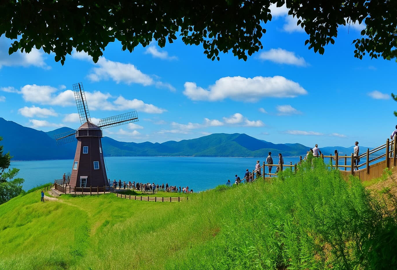 Tourists walking along a scenic coastal path beside a windmill at Windy Hill in Geoje, South Korea, overlooking the ocean and surrounding mountains.