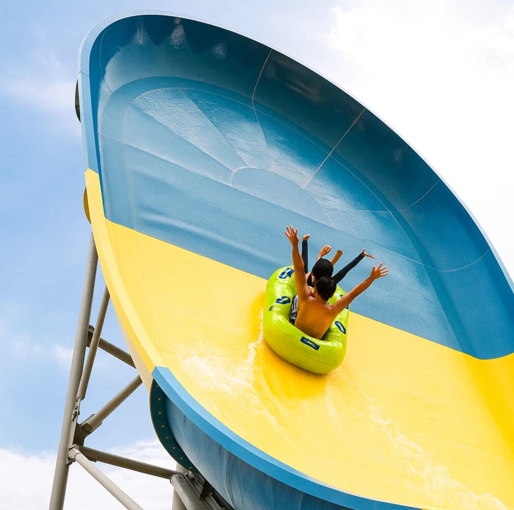 Two riders in a green inflatable raft enjoying a high-speed descent on a large yellow and blue water slide at a Geoje waterpark under a sunny sky.